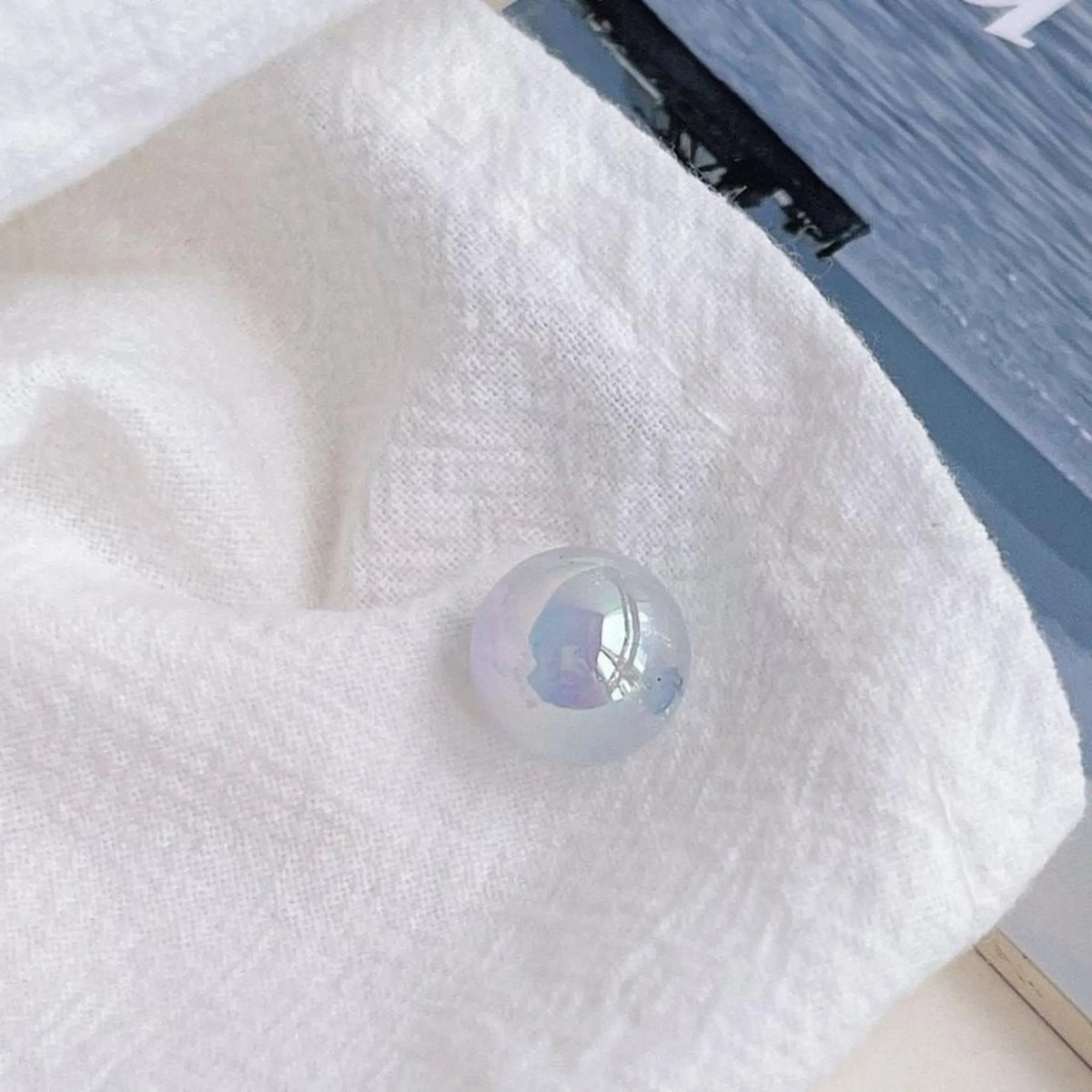 A pale blue acrylic round bead with an iridescent finish shown on textured white cloth in daylight.