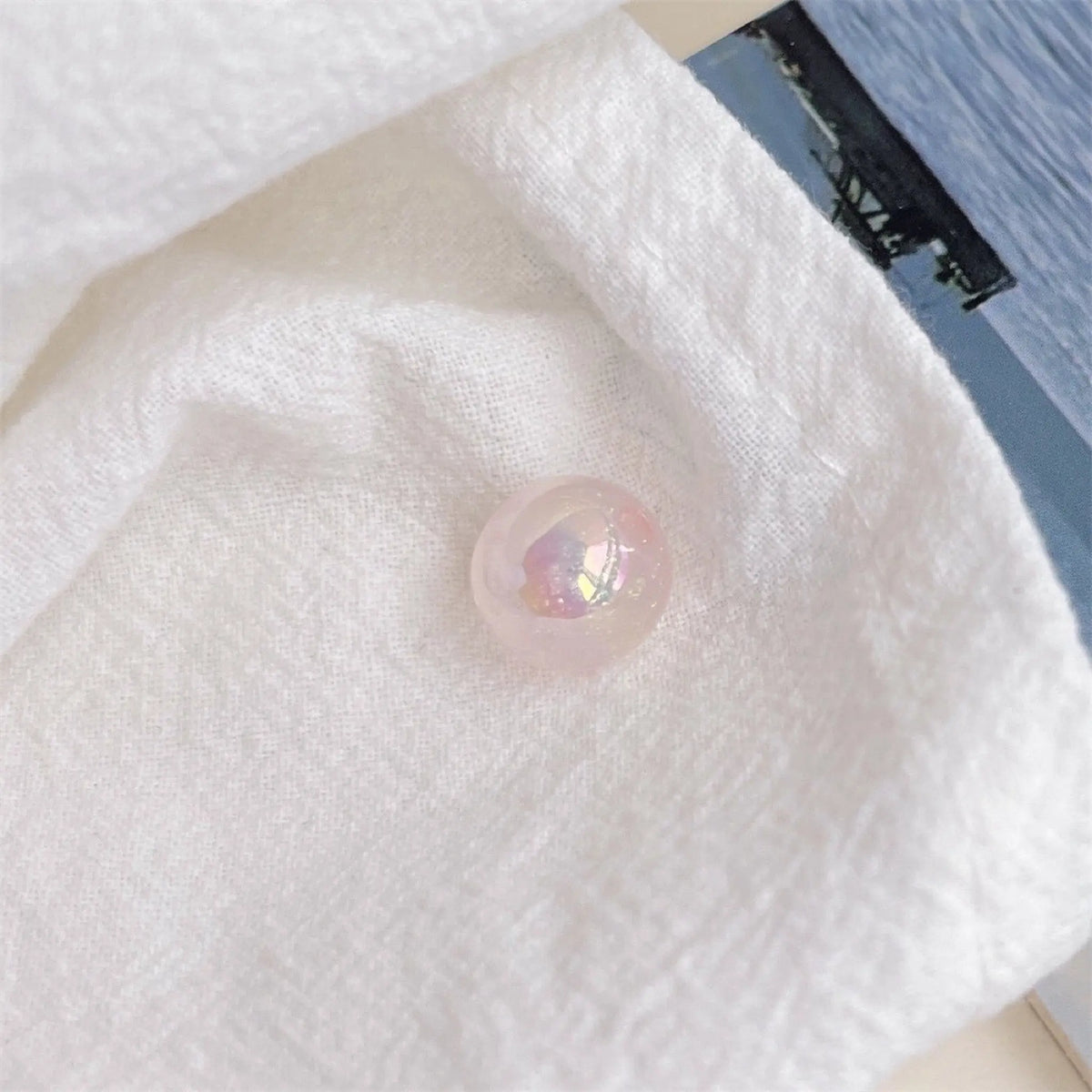 A pale pink acrylic round bead with an iridescent finish shown on textured white cloth in daylight.