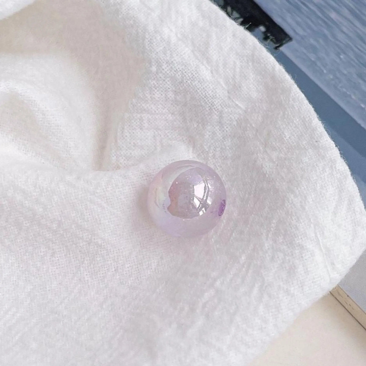 A pale purple acrylic round bead with an iridescent finish shown on textured white cloth in daylight.