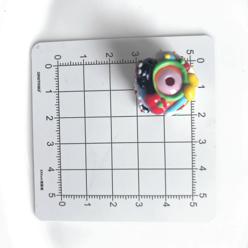 Hole view of one decorated acrylic bead on a centimeter grid, showing the threading hole and raised colored details.