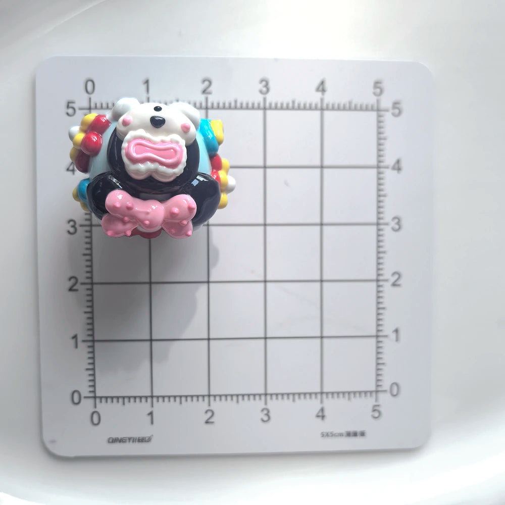 Front view of one decorated acrylic bead with a pink bow on a centimeter grid for size reference.