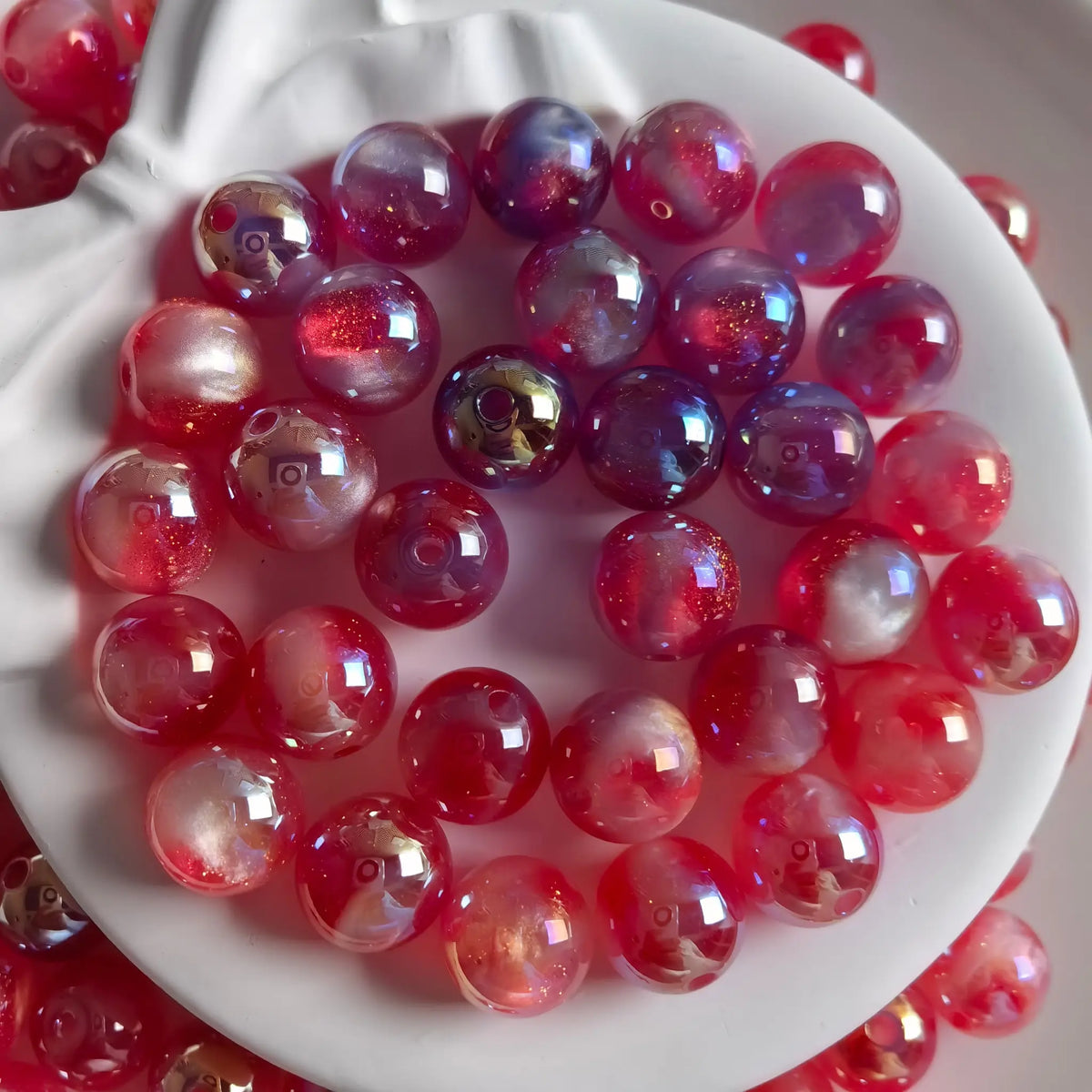 Red iridescent acrylic round beads with glossy reflections and fine shimmer in a white bow-shaped dish.