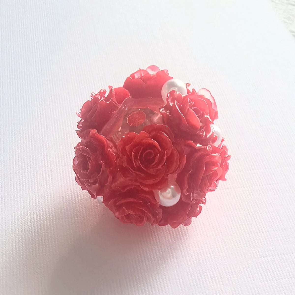 Red rose flower ball bead with raised rose petals, pearl-like accents and a threading hole, shown on white fabric.